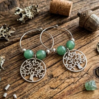 une paire de boucles d'oreilles en argent avec sur chacune deux perles d'aventurine verte et un arbre de vie posées sur une table en bois