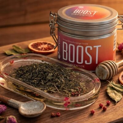 Une boite de thé de la marque Alveus qui s'appelle Boost avec une coupelle remplie de feuilles de thé posées sur une table en bois