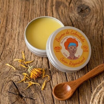 Un baume de calendula de couleur jaune avec son couvercle couvert d'un dessin avec un visage de femme et un texte qui dit baume de calendula, soin régénérant, posé sur un fond en bois avec une fleur jaune et une cuillère en bois et un morceau de corde
