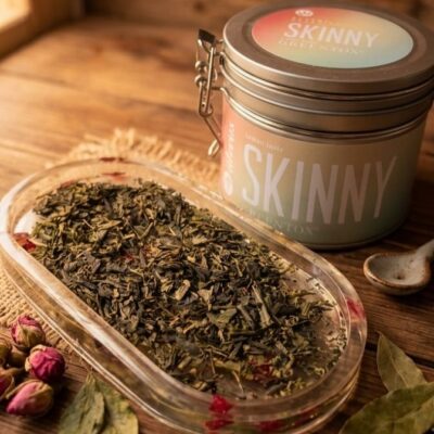 Une boite de thé de la marque Alveus qui s'appelle Skinny avec une coupelle remplie de feuilles de thé posées sur une table en bois