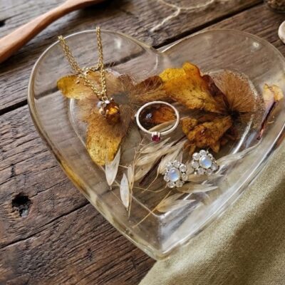 Des bijoux, bague, collier, boucle d'oreilles sur une coupelle en forme de cœur en résine époxy avec des inclusions de fleurs séchées. Posé sur une table en bois