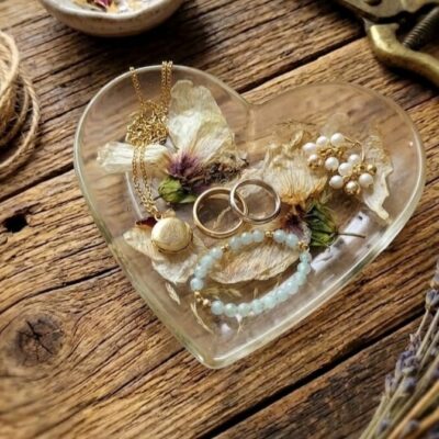 Des bijoux sur une coupelle en forme de cœur en résine époxy avec des inclusions de fleurs séchées. Posé sur une table en bois