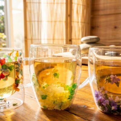 3 tasses en verre transparent avec chacune des inclusions de fleurs colorées et remplies de thé. Posées sur une table en bois
