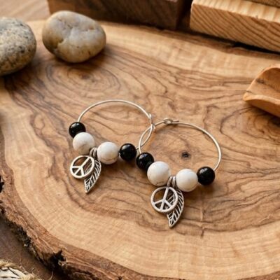 Une paire de boucles d'oreilles rondes avec deux pierres blanches et deux pierres noires ainsi que é breloques , une feuille et un symbole de paix. Posées sur une planche en bois entourées de cailloux gris