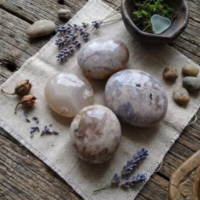 quatre pierres d'agate fleur de cerisier aux tons beiges posées sur un carré de jute sur une table en bois, entourées de fleurs de lavande, de petits cailloux et d'un petit bol en terre avec de la mousse verte a l'intérieur ainsi qu'une pierre en forme de cœur transparente.