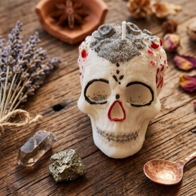 Une bougie en cire de soja en forme de crane calavera de couleurs blanche, noire et rouge posée sur une table en bois et entourée de fleurs de lavande, de pierres dorées , de pétales rouges et d'une cuillère en métal argenté.