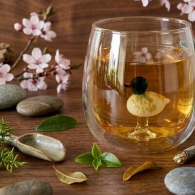 Une tasse en verre transparent avec une ance verte transparent et a l'intérieur de la tasse une figurine en forme de canard vert et blanc. Posée sur une table en bois et entourée de fleurs de cerisier, cailloux, feuilles vertes