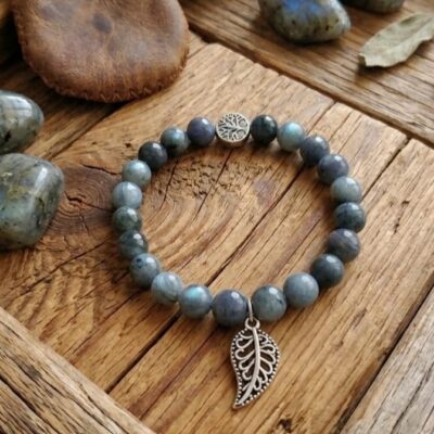 Un bracelet en perles de labradorite grises avec une breloque qui représente une feuille et un cache nœud qui représente un arbre de vie. Posé sur un fond en bois et entouré de pierres de labradorite et d'un pochon en cuir marron