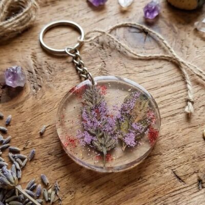Un porte clefs en résine époxy avec des inclusions de fleurs vertes et des paillettes rouges et violettes, posé sur une table en bois et entouré de fleurs de lavande, d'un morceau de corde et de pierres violettes