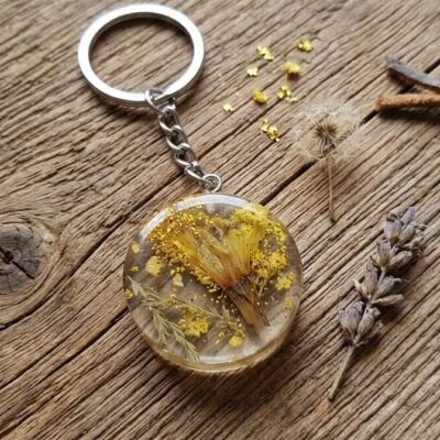 Un porte clefs en résine époxy avec des inclusions de fleurs naturelles jaunes et vertes, posé sur un fond en bois et entouré d'une fleur de lavande , d'un pissenlit et d'un morceau de cuir marron