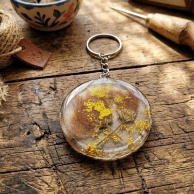 Un porte clef de forme ronde en résine époxy avec des inclusions de fleurs séchées marrons et des paillettes jaunes. Posé sur une table en bois et entouré de fleurs de pissenlit et d'un rouleau de corde