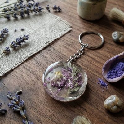 Un porte clef de forme ronde en résine époxy avec des inclusions de fleurs séchées vertes et violettes et des paillettes violettes . Posé sur une table en bois et entouré de fleurs de lavande, d'un carré de toile de jute et de petits cailloux marrons.