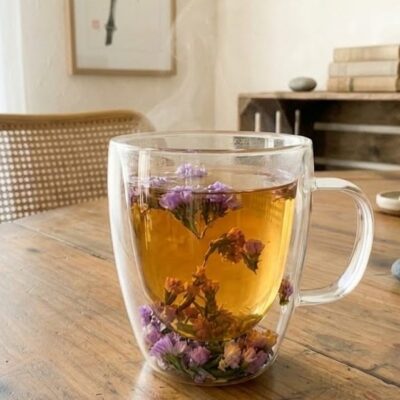 Une tasse en verre transparent avec des inclusions de fleurs violettes dans la paroi remplie de thé posée sur une table en bois